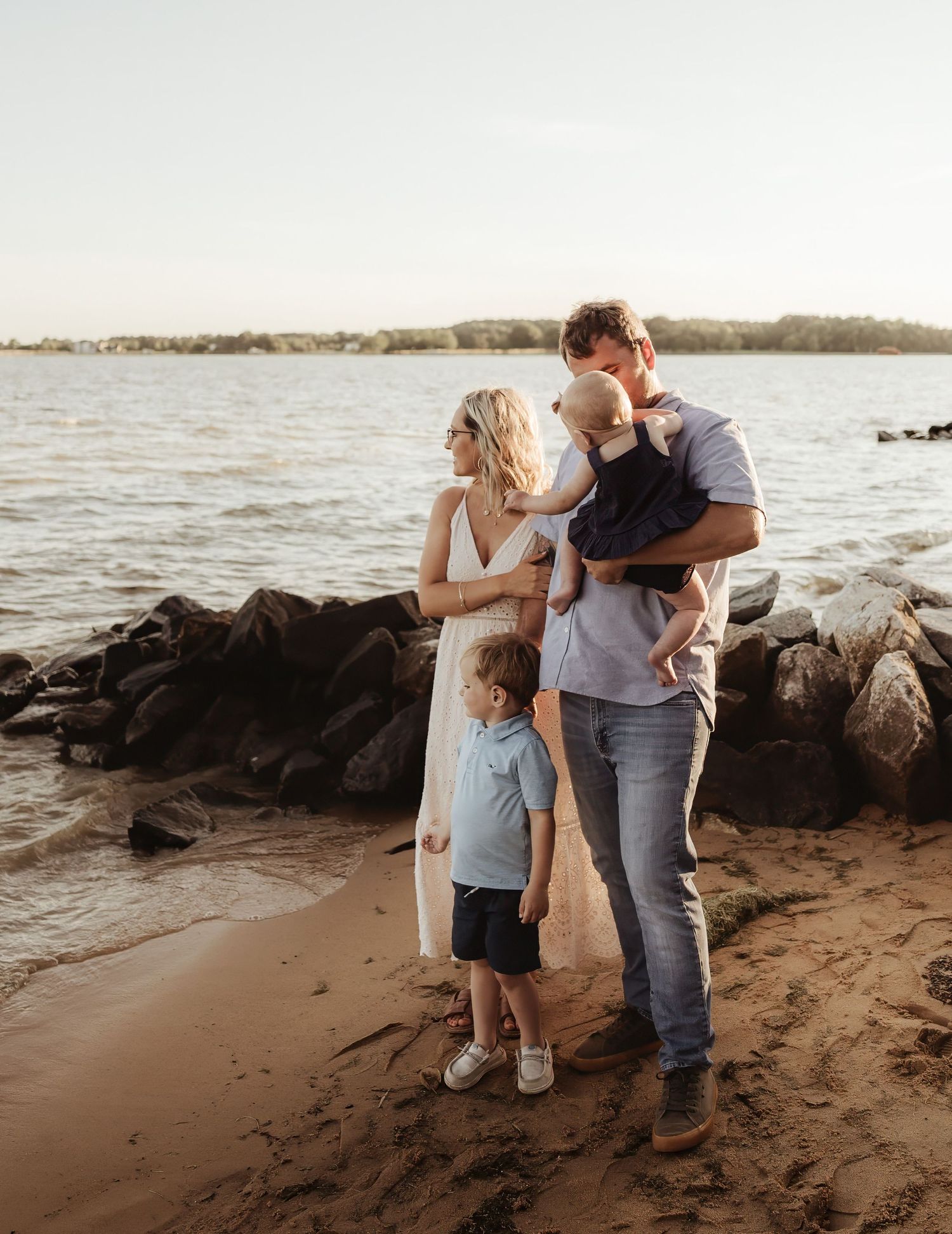 Family of four standing on sandy beach by rocky shore, parents holding toddler while young boy stands nearby.