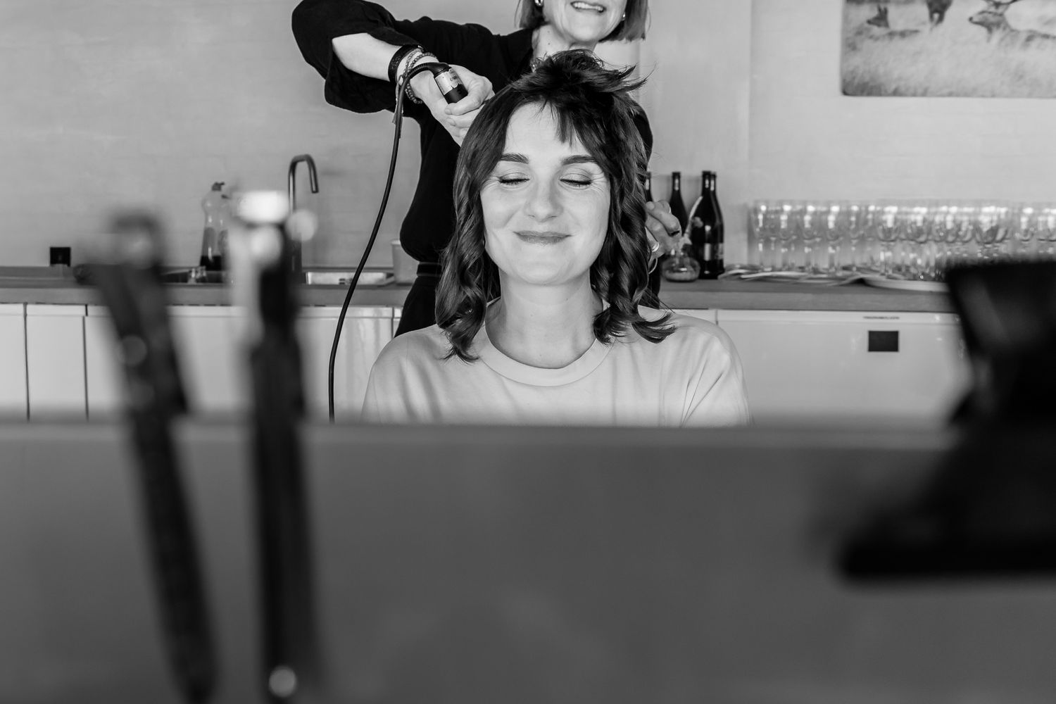 Black and white photo of a smiling woman getting her hair styled by a hairdresser in a bright salon.