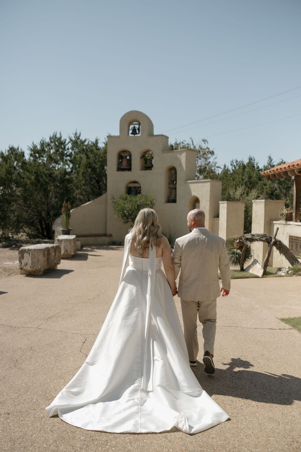 Bride and groom walking hand in hand toward Spanish-style chapel with bell tower.