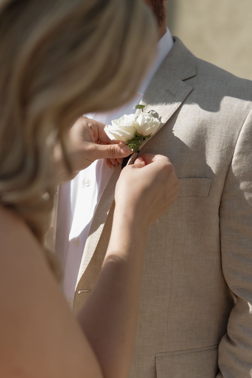 Bride pinning white rose boutonniere on groom wearing beige linen wedding suit.