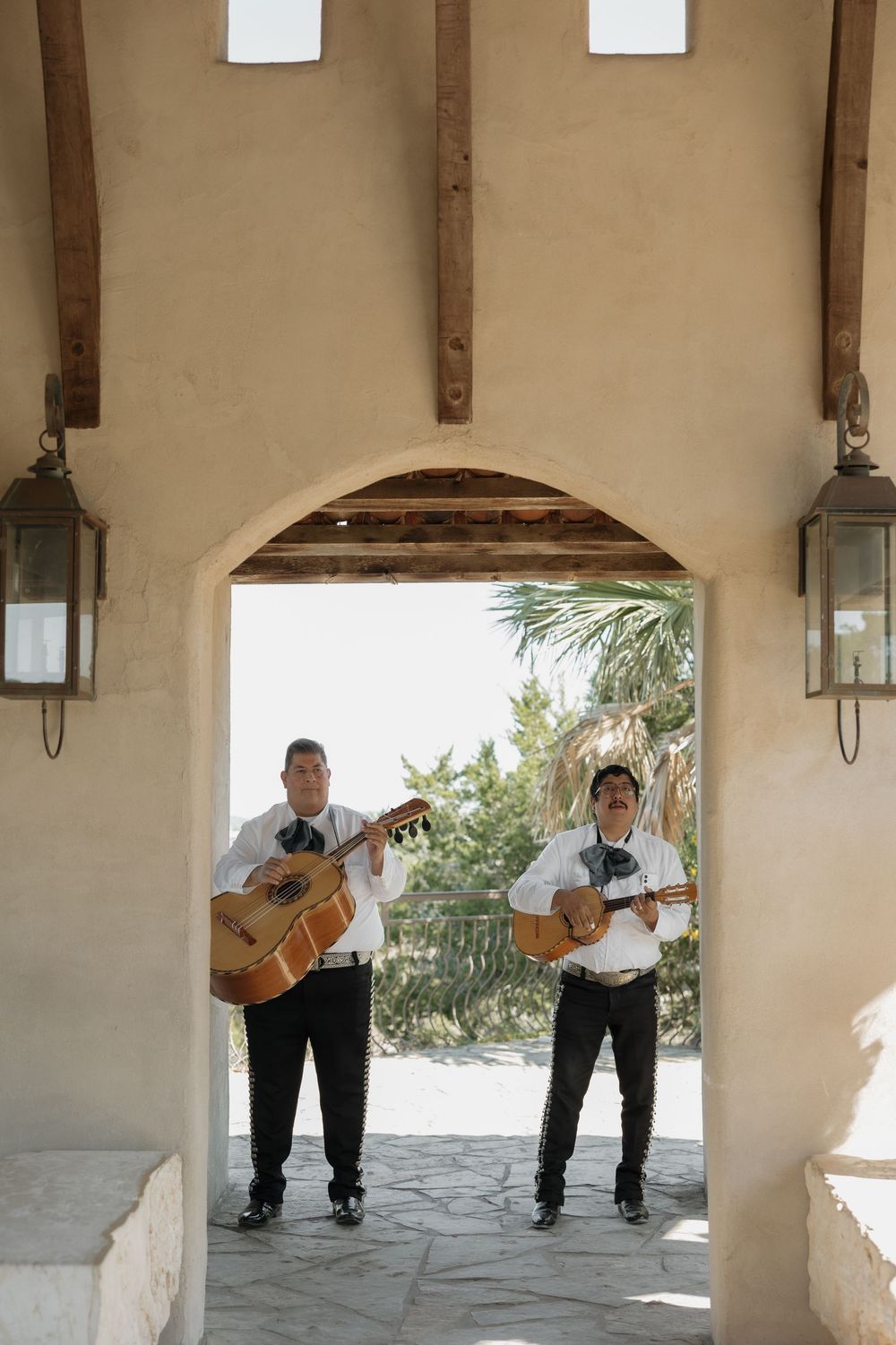 Two mariachi musicians playing guitars under arched doorway with lanterns at wedding venue.