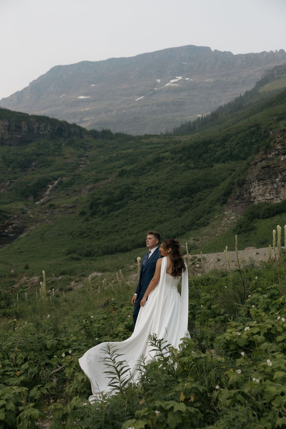 Bride and groom stand in a lush mountain meadow, her white gown flowing in the wind, with rocky peaks behind.