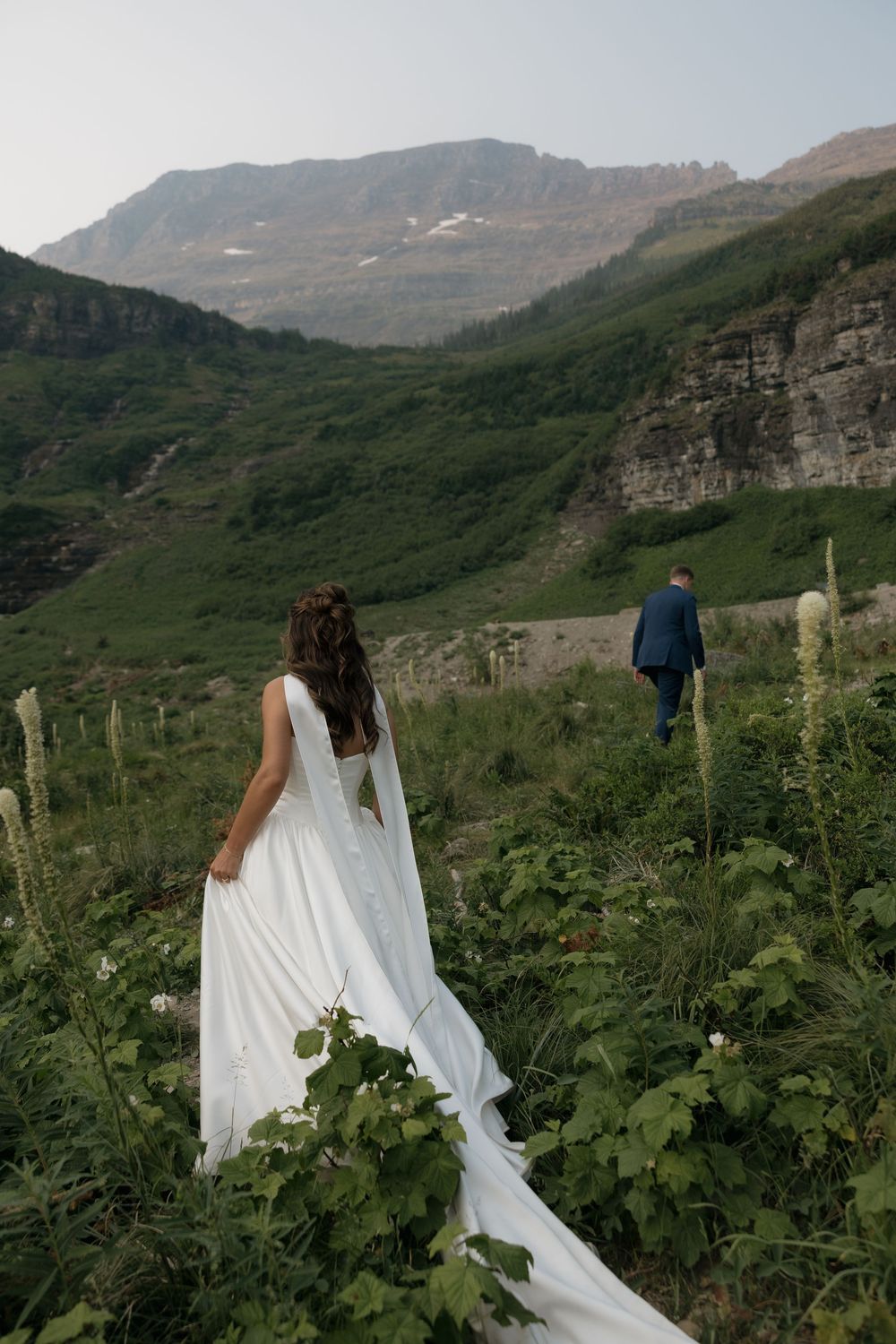 Bride in white gown walks through wildflowers toward groom in blue suit with mountain backdrop.