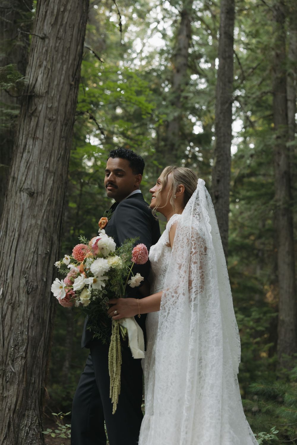 Bride in lace gown and veil holds floral bouquet beside groom in black suit among tall forest trees.