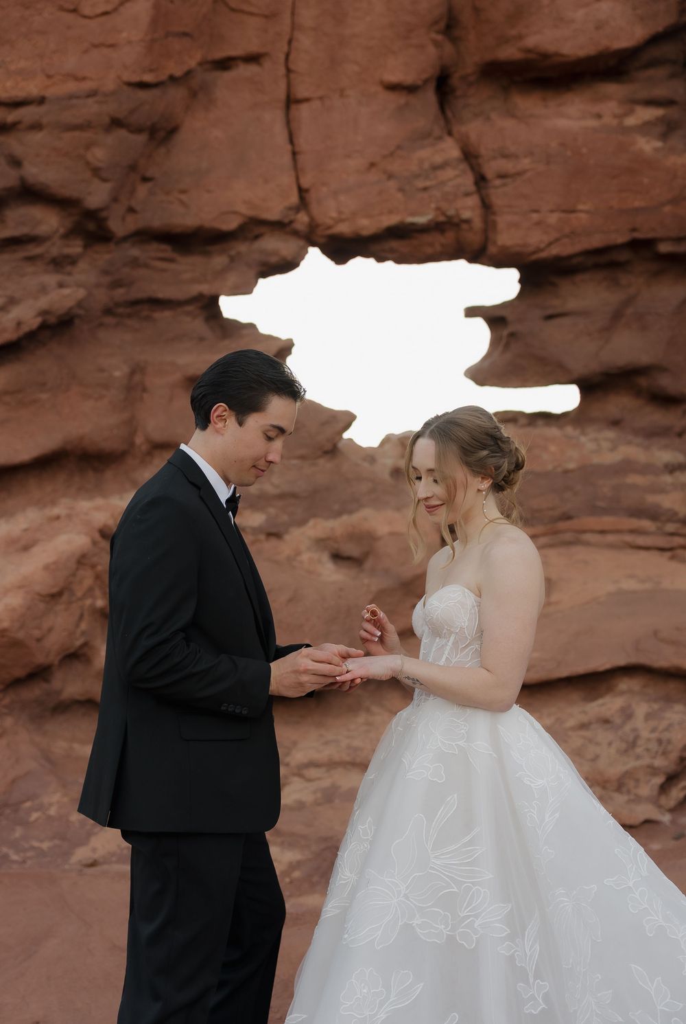 Groom places wedding ring on bride's finger before a natural red rock arch in an outdoor ceremony.