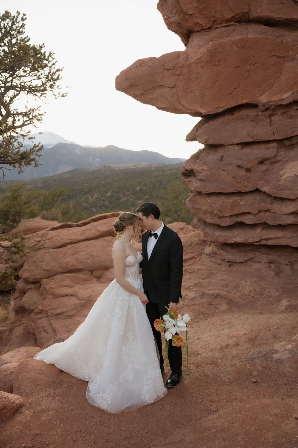 Wedding couple shares a tender moment beside red rock spires with mountain views at dusk.