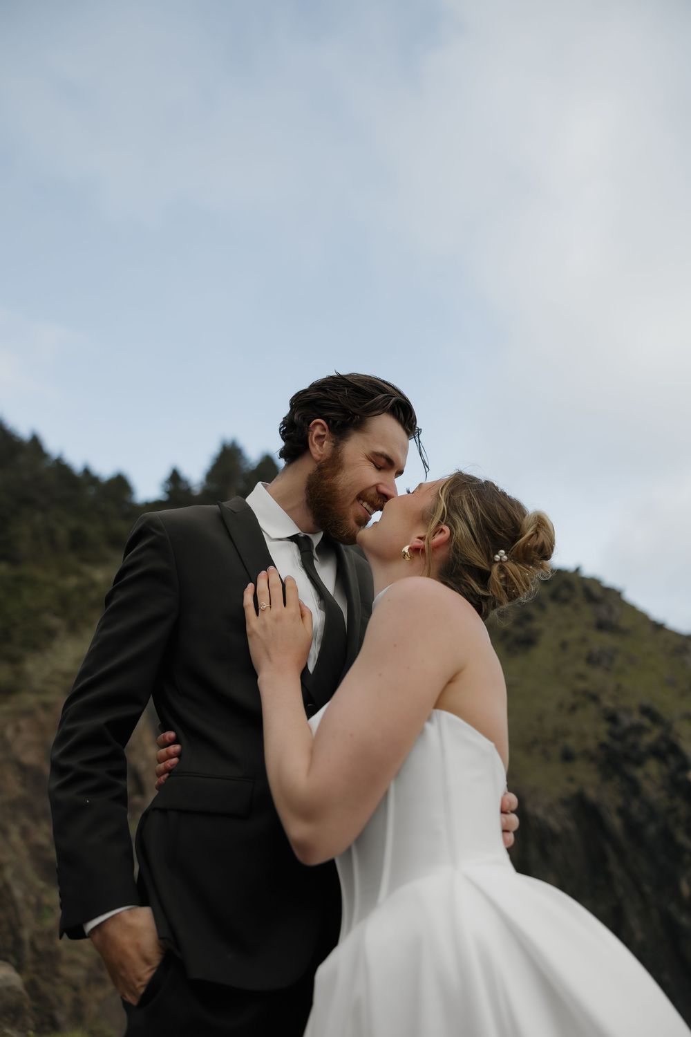 A smiling bride and groom share an intimate moment about to kiss with green mountains behind them.