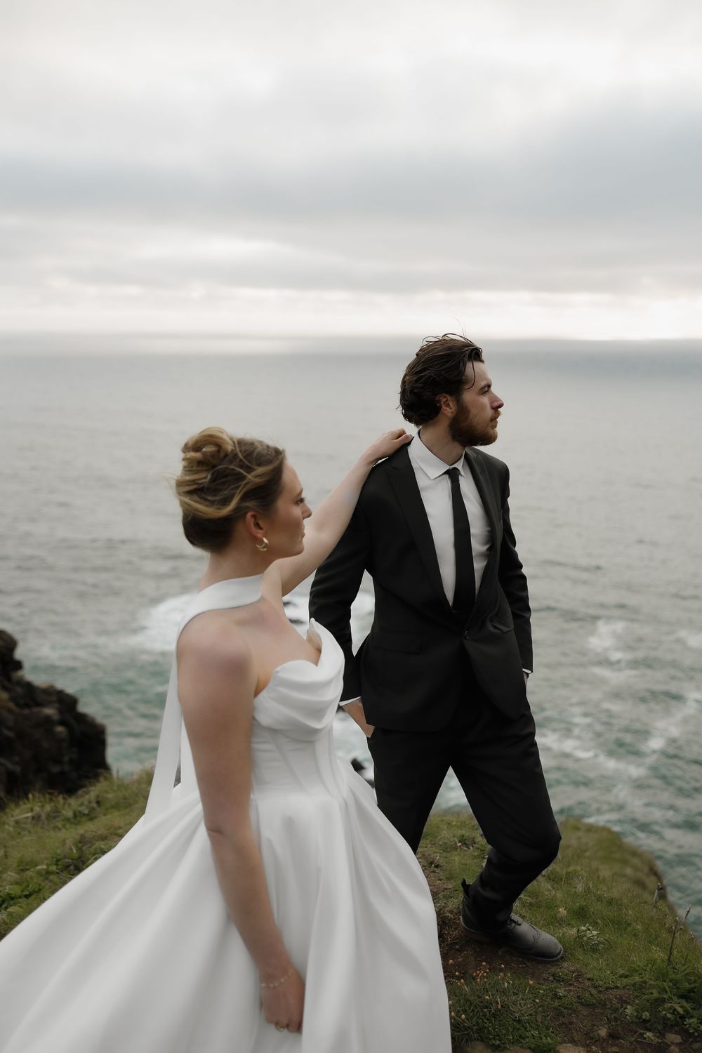 A bride in a strapless white gown and groom in a black suit stand on a scenic ocean overlook.
