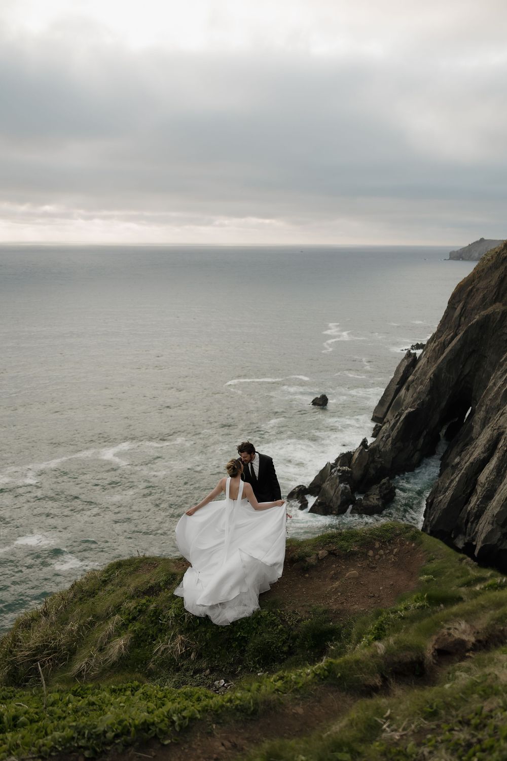 A bride and groom share a kiss on a dramatic coastal cliff with crashing waves and rocks below.