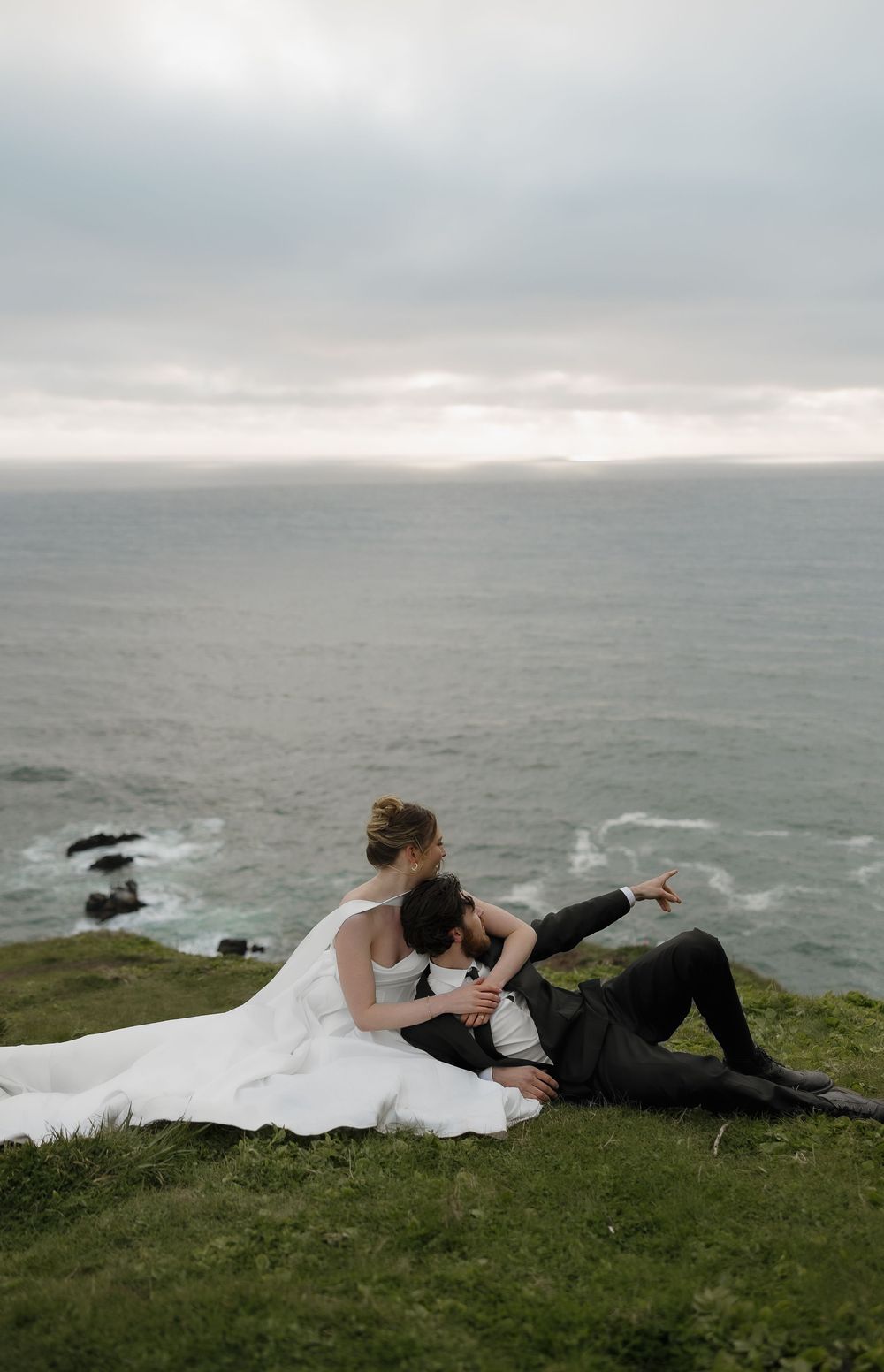 A bride in white and groom in black relax together on a grassy ocean cliff in a romantic pose.