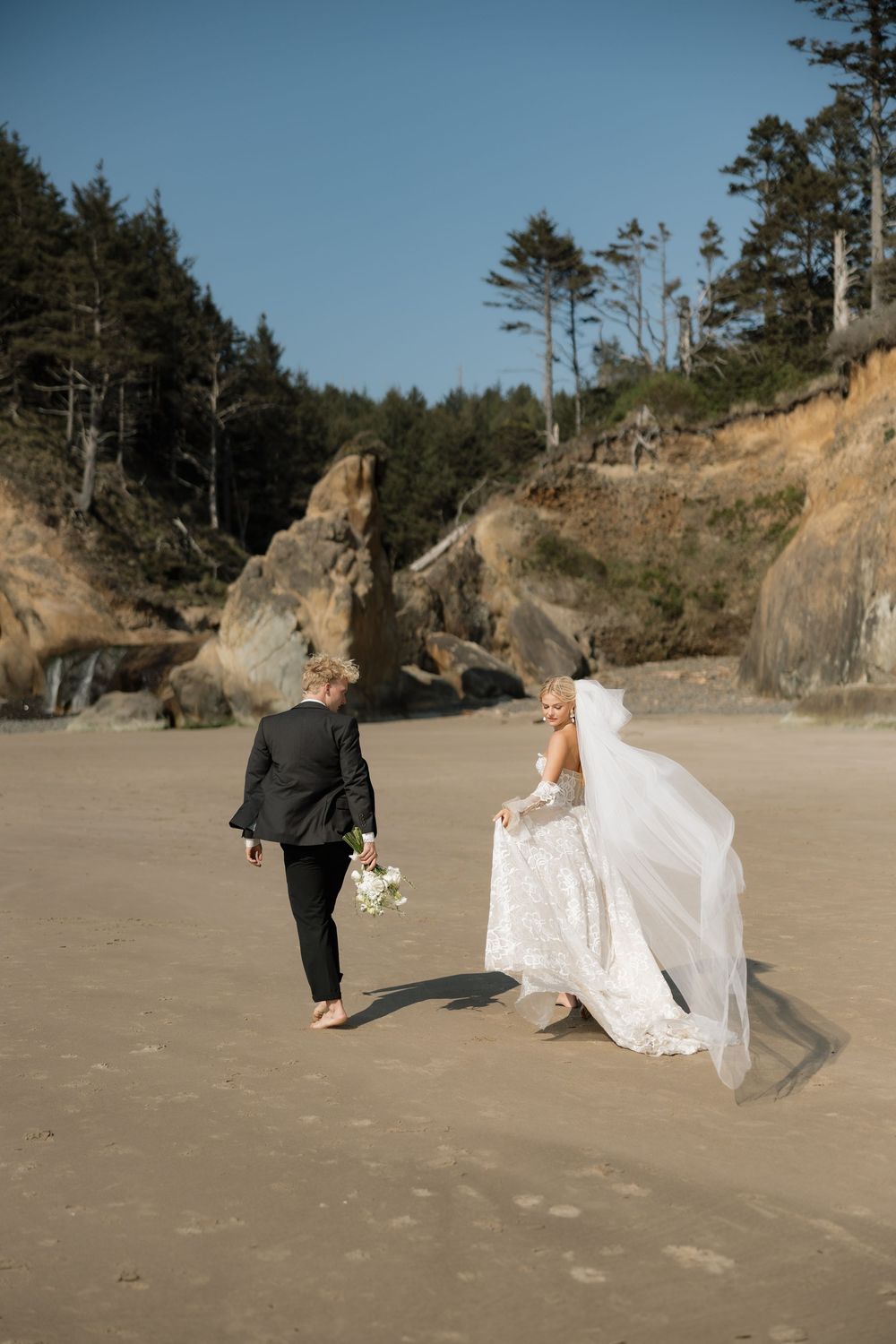 Bride in flowing lace gown with veil blowing in wind walks toward groom on sandy beach with rocky cliffs.