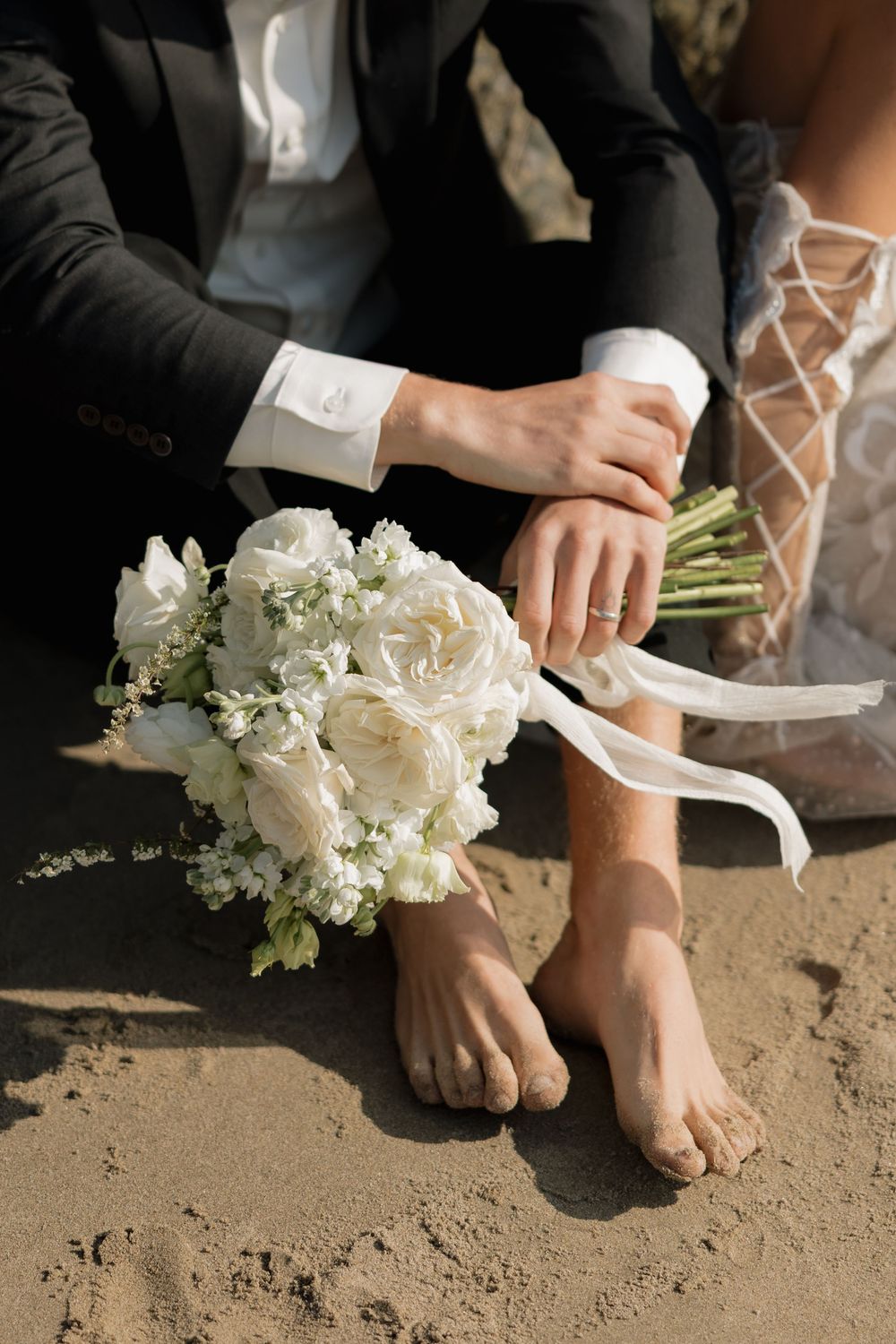 Close-up of bride and groom sitting barefoot in sand with a white rose wedding bouquet tied with ribbon.