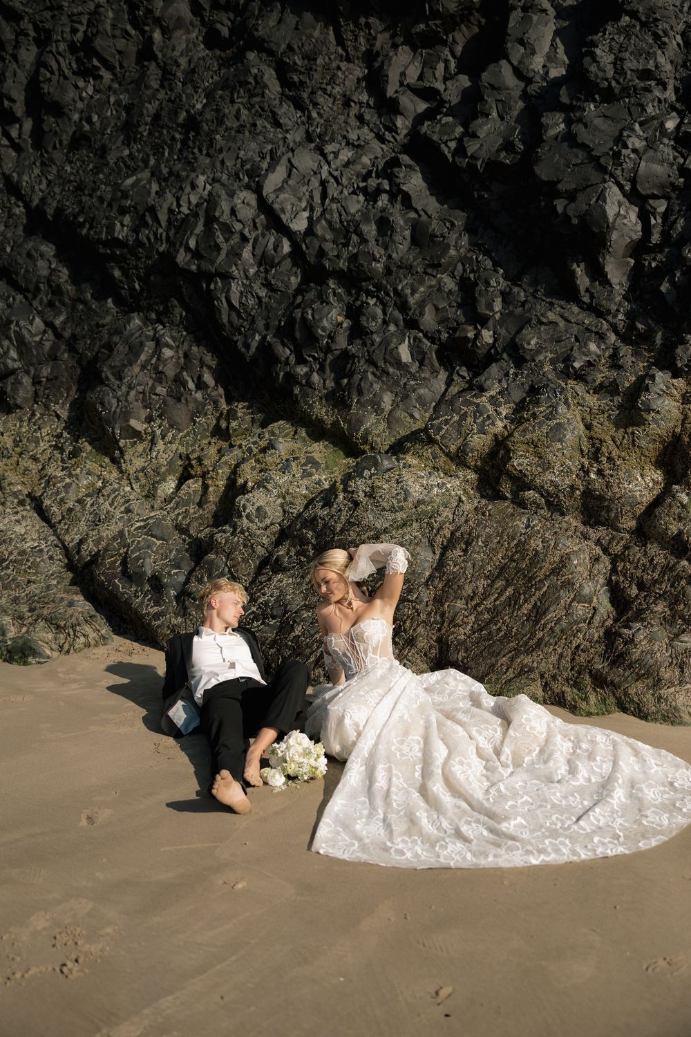 Bride and groom sit barefoot on sandy beach leaning against dark mossy coastal rock cliffs.
