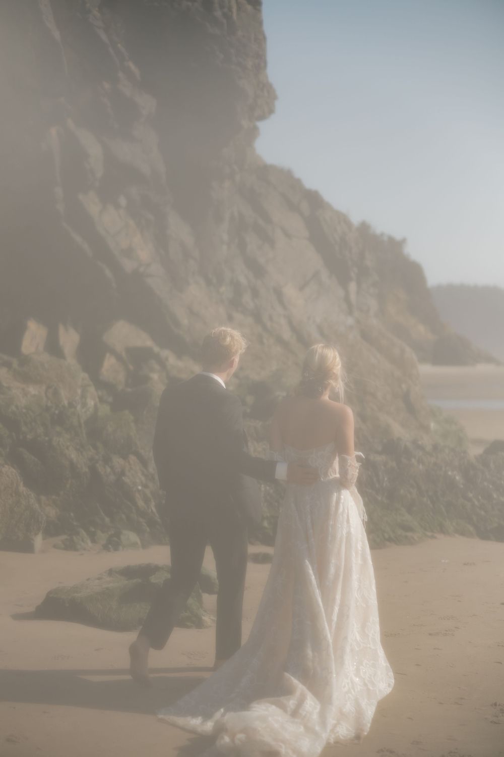 Bride and groom walk hand in hand along a misty sandy beach beside dramatic coastal cliffs.