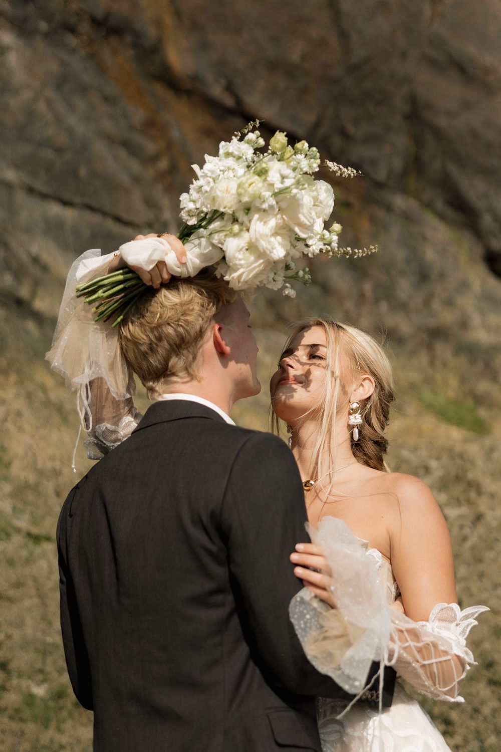 Laughing bride tosses bouquet while embracing groom against a rocky outdoor backdrop in sunlight.