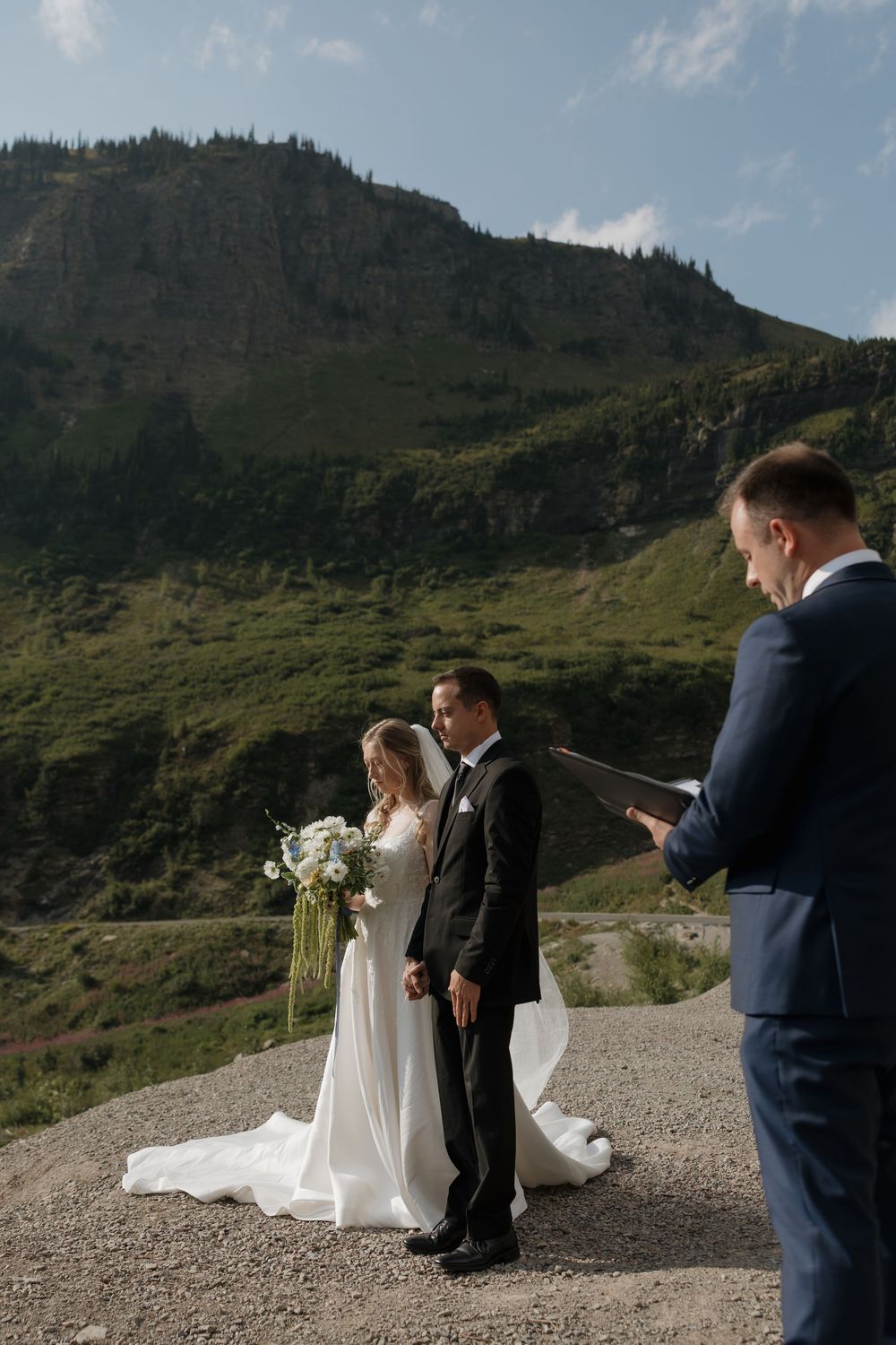 Couple exchanges vows at outdoor mountain wedding ceremony with officiant, bride holding white bouquet, scenic green hills backdrop.