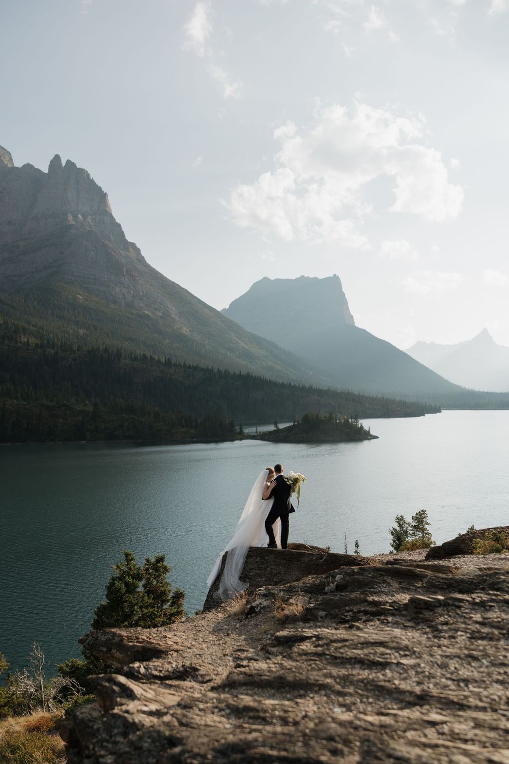 Bride and groom embrace on a rocky cliff overlooking a serene mountain lake with misty peaks in the background.