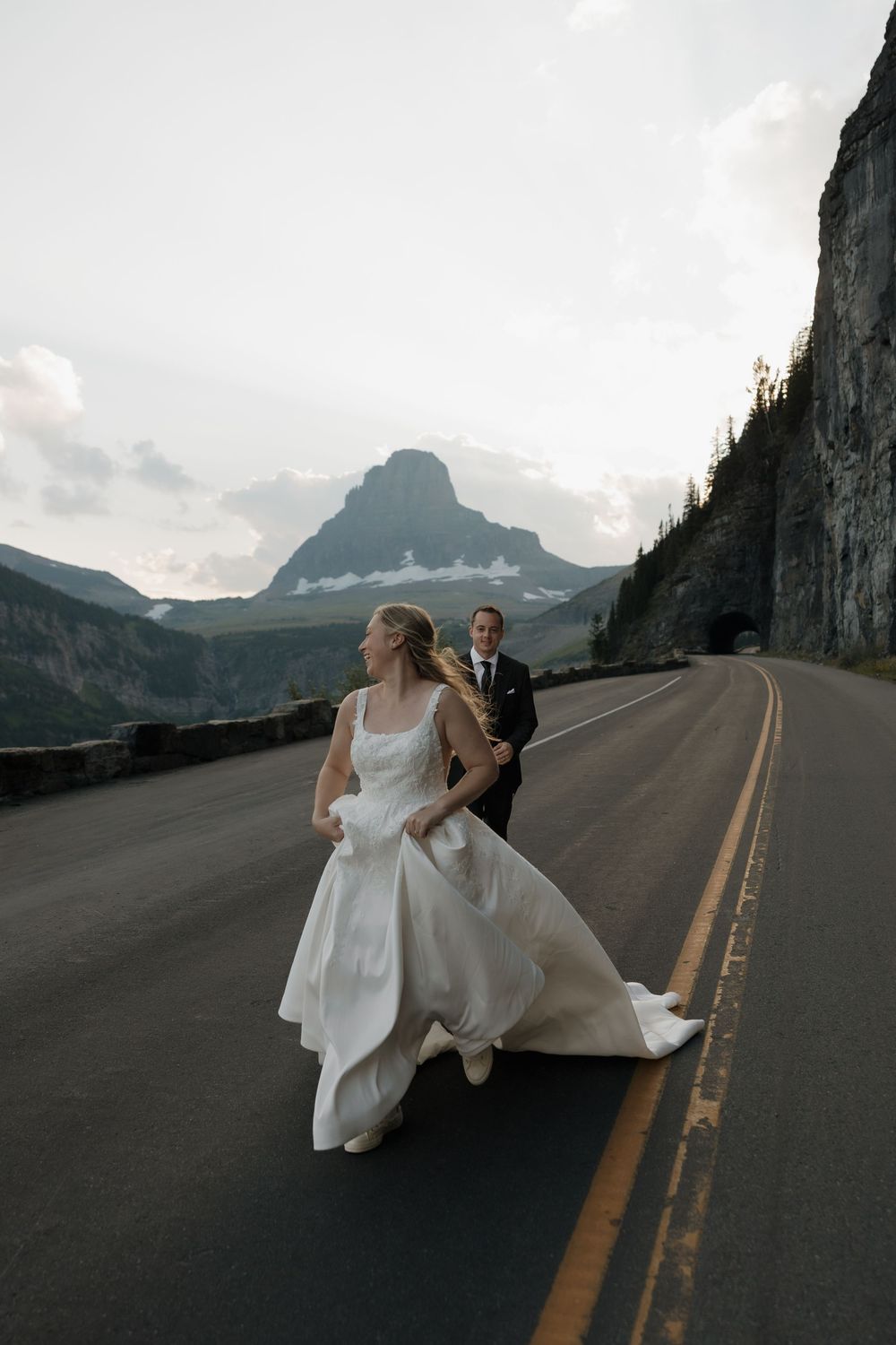 Bride and groom walk on mountain road in Glacier National Park, white wedding gown flowing, dramatic peaks behind them.