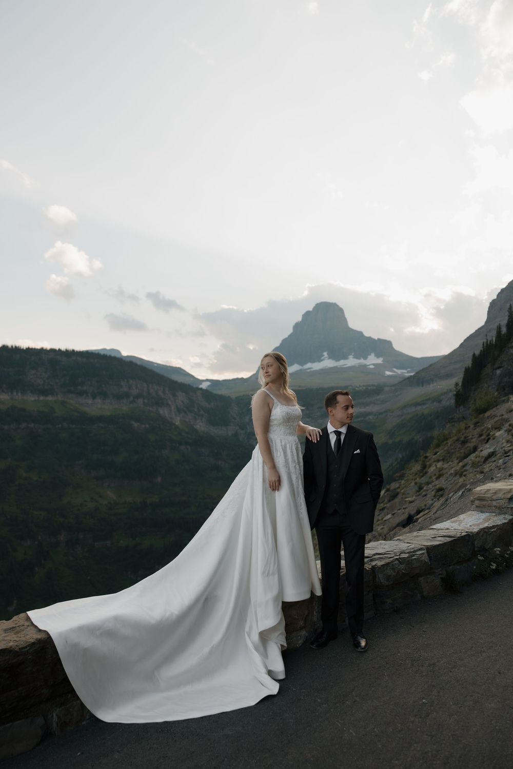 Bride and groom pose on a mountain overlook in Glacier National Park, with dramatic peaks and clouds behind them.