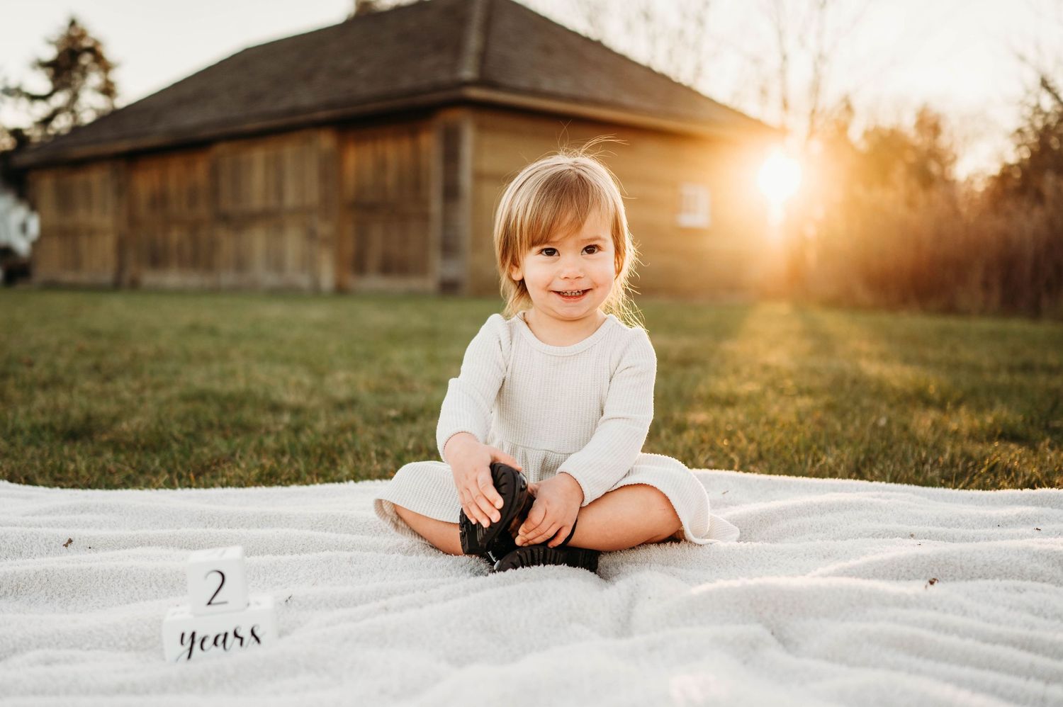 A smiling toddler sits on a white blanket outdoors at golden hour, wearing a white onesie, with a rustic barn behind.