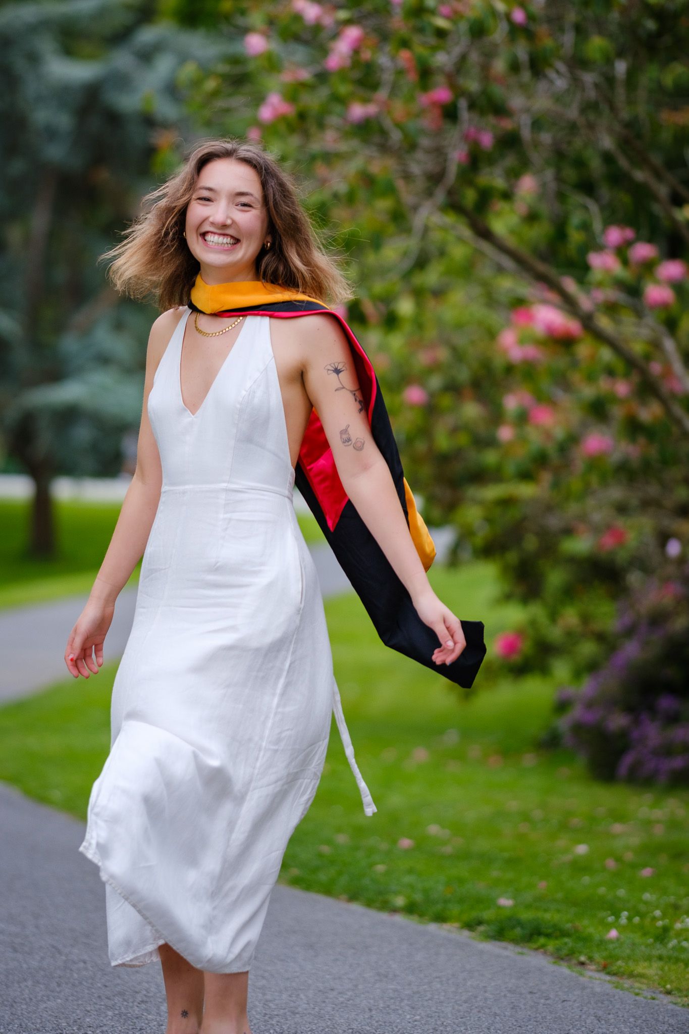 Smiling young woman in white dress with graduation stole walks through a blooming garden park.