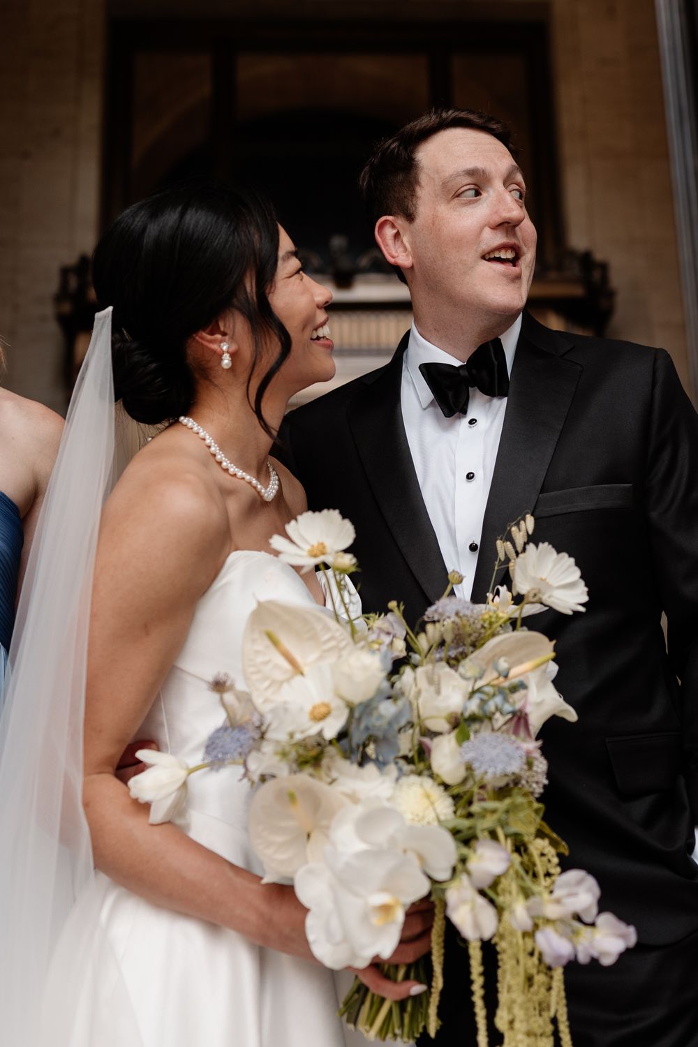 Bride and groom in black tuxedo smiling together holding white floral bouquet on wedding day.
