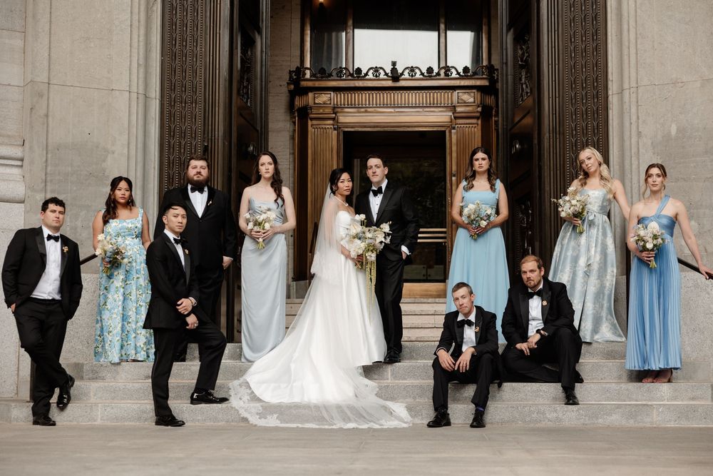 Wedding party poses on grand steps, bride in white gown with groomsmen in tuxedos and bridesmaids in blue dresses.