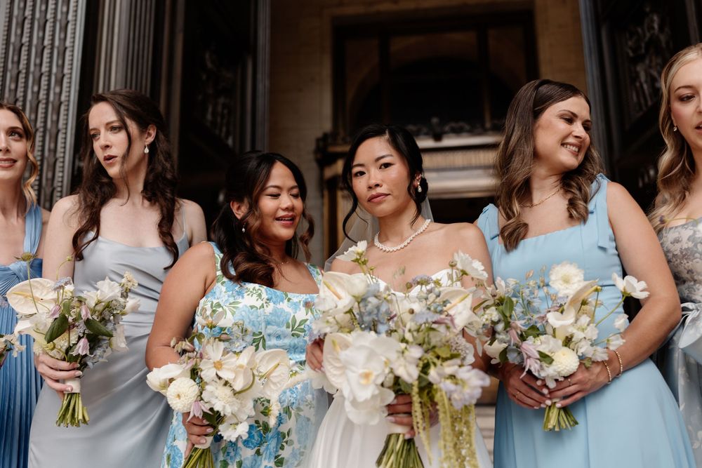 Bride in white gown with bridesmaids in blue dresses holding white and blue floral bouquets at a wedding ceremony.