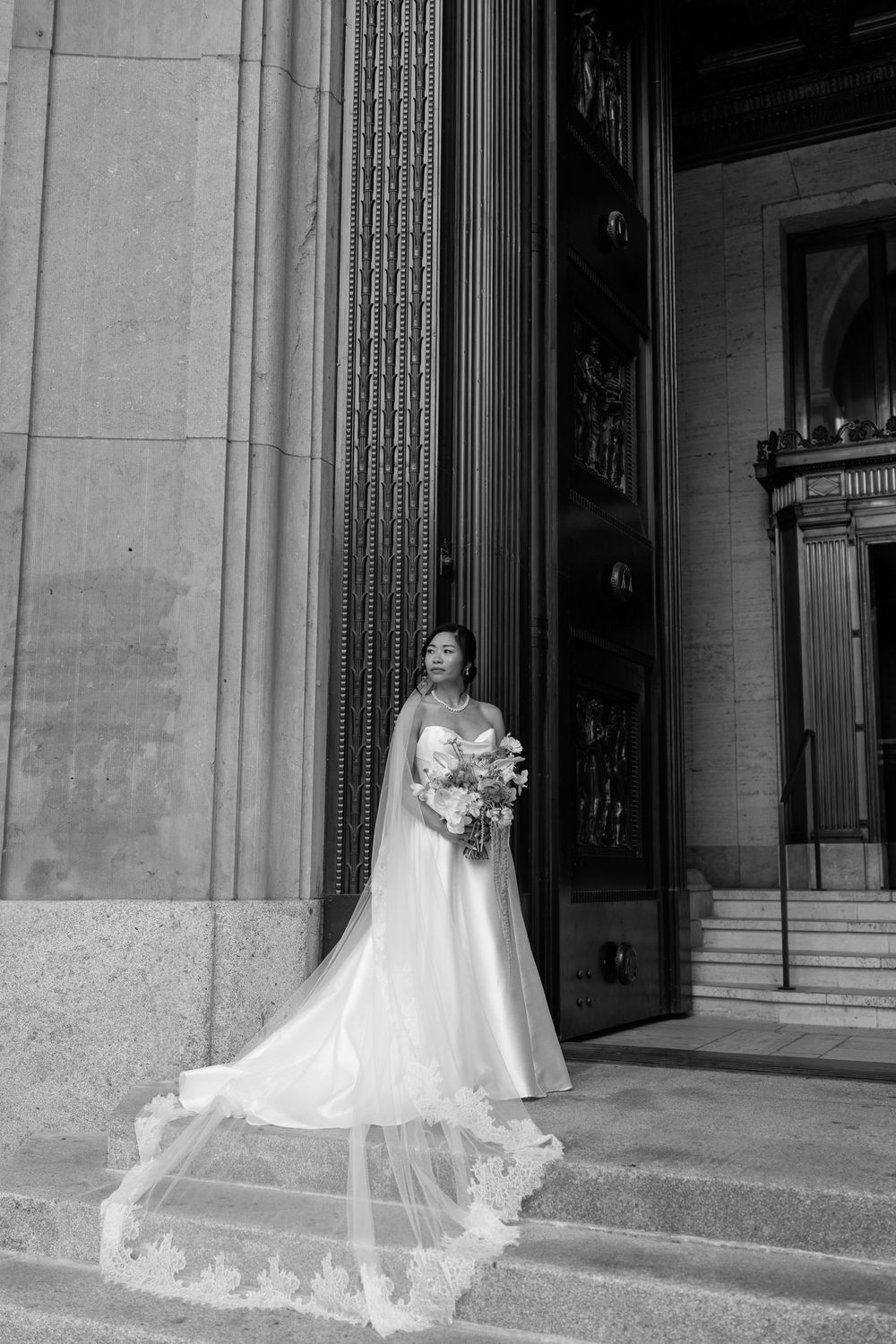 Bride in strapless gown with lace-trimmed cathedral veil stands on steps of grand stone building holding bouquet.