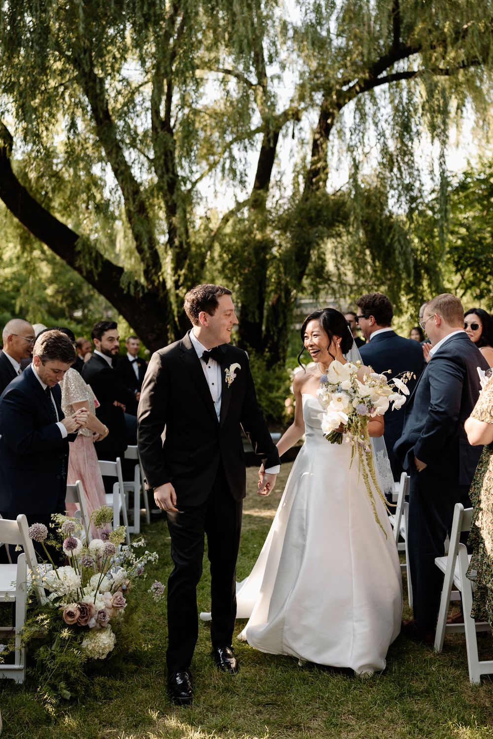 Newlywed couple walks down outdoor aisle under willow tree surrounded by wedding guests.