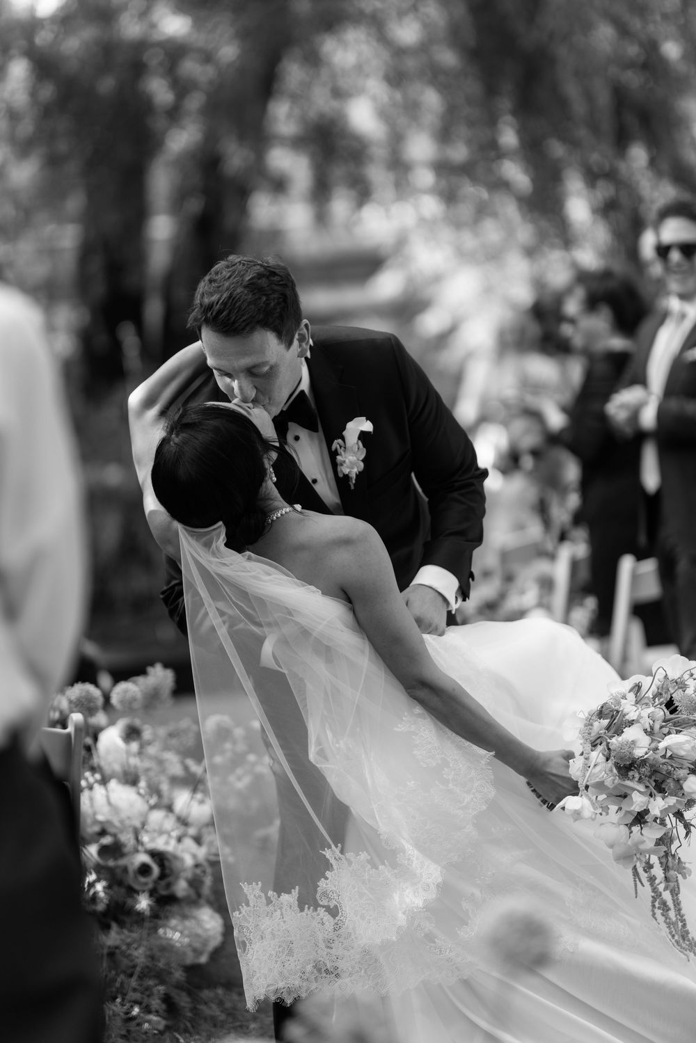 Groom dips bride for a romantic kiss at outdoor wedding ceremony in black and white photography.