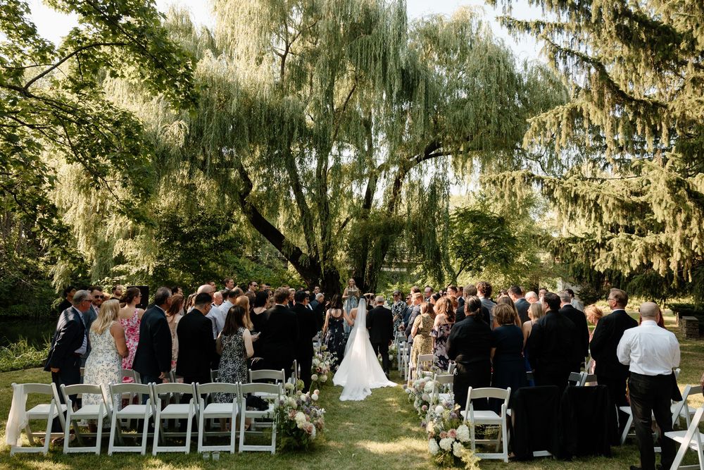 Outdoor wedding ceremony under weeping willow tree with guests seated on white chairs.