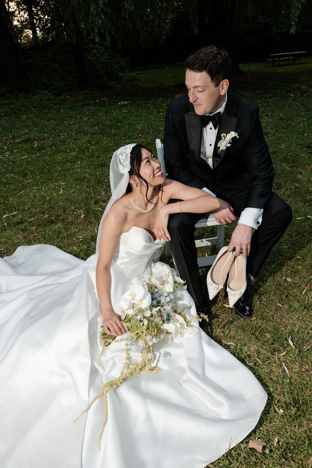 Bride in white gown with bouquet sits on grass as groom in tuxedo holds her heels in a garden setting.