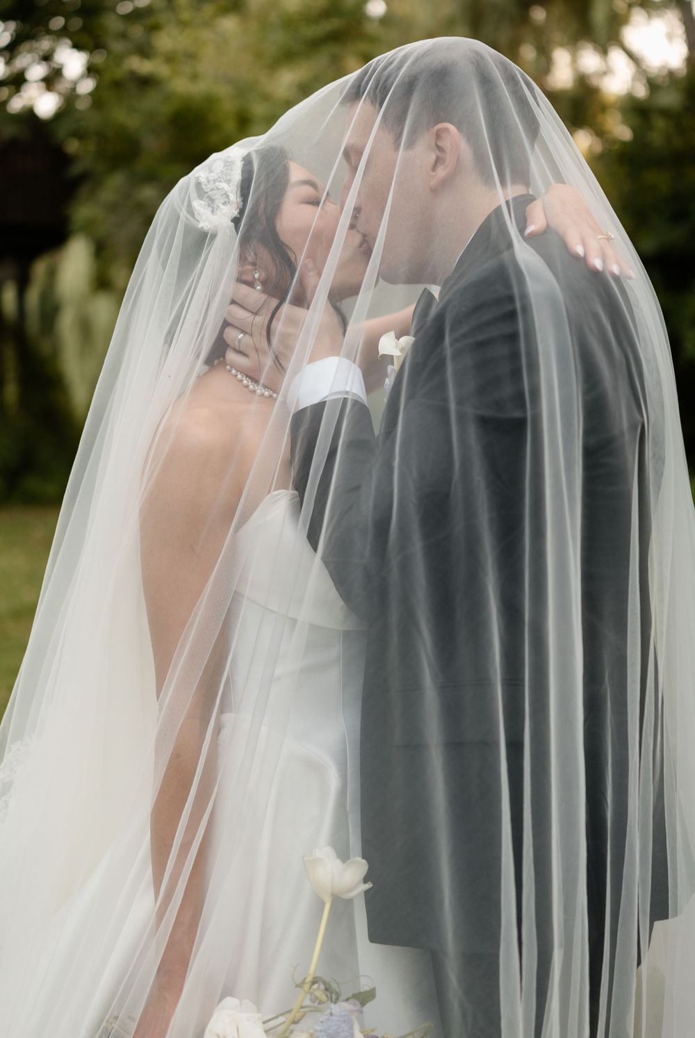 Bride and groom share a romantic kiss hidden beneath a flowing white veil outdoors on their wedding day.