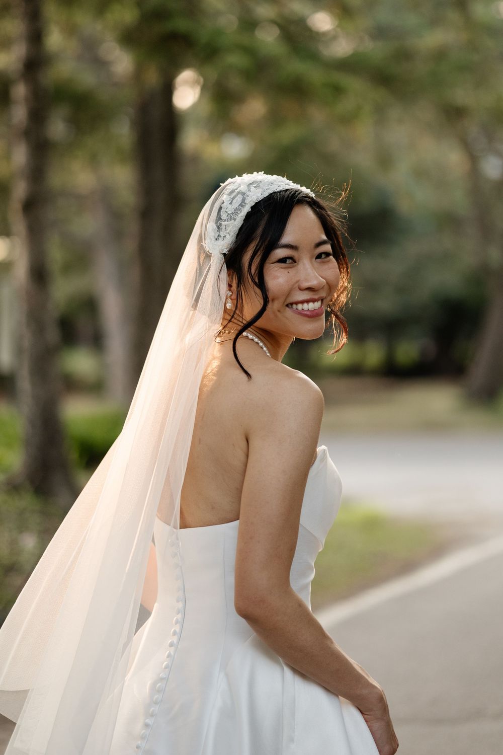 Smiling bride in strapless white gown and lace veil poses outdoors among trees on her wedding day.