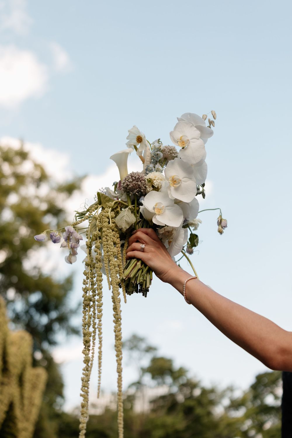 Hand holding elegant white orchid and wildflower wedding bouquet against blue sky outdoors.