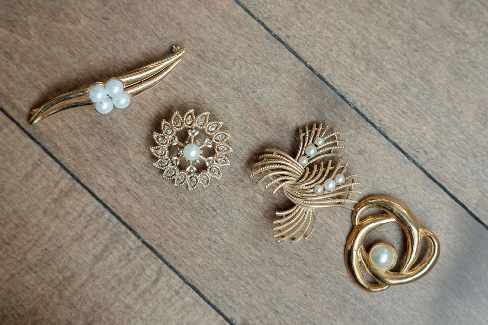 Four vintage gold and pearl brooches arranged on rustic wood surface, featuring floral and abstract designs.