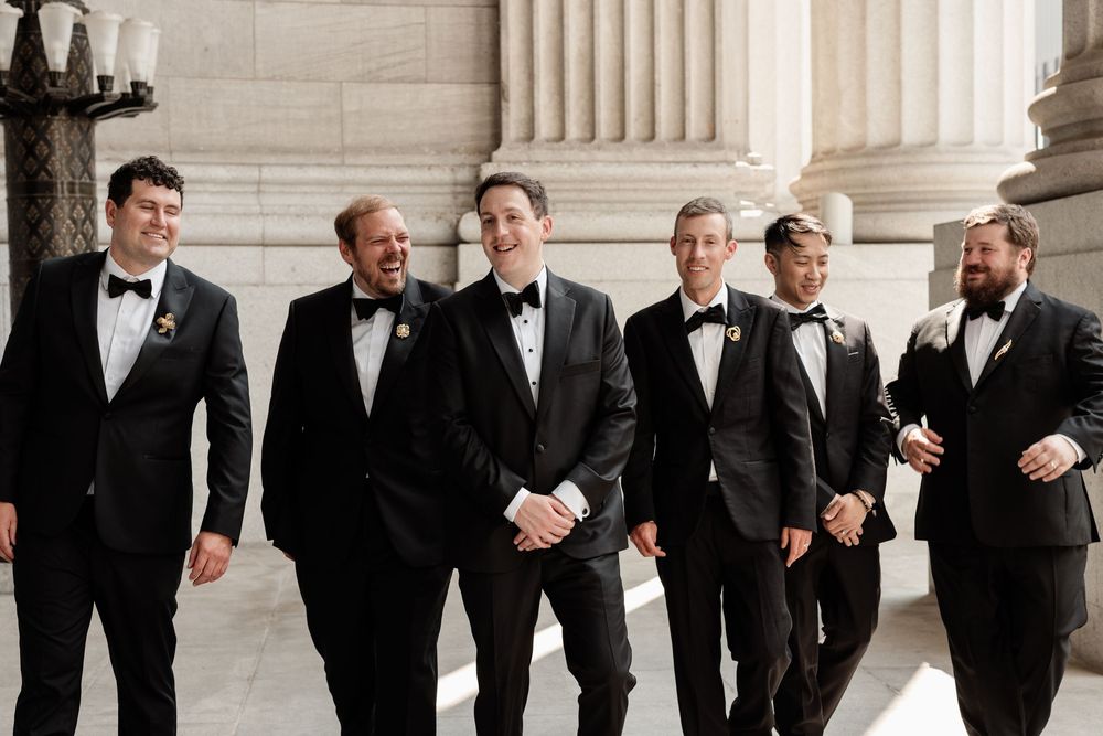 Six groomsmen in black tuxedos with bow ties laugh and walk together outside elegant stone columns.
