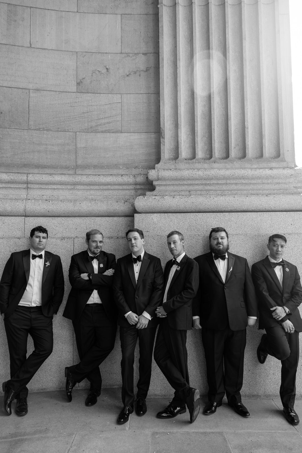 Six groomsmen in black tuxedos leaning against a stone column in a classic black and white wedding photo.