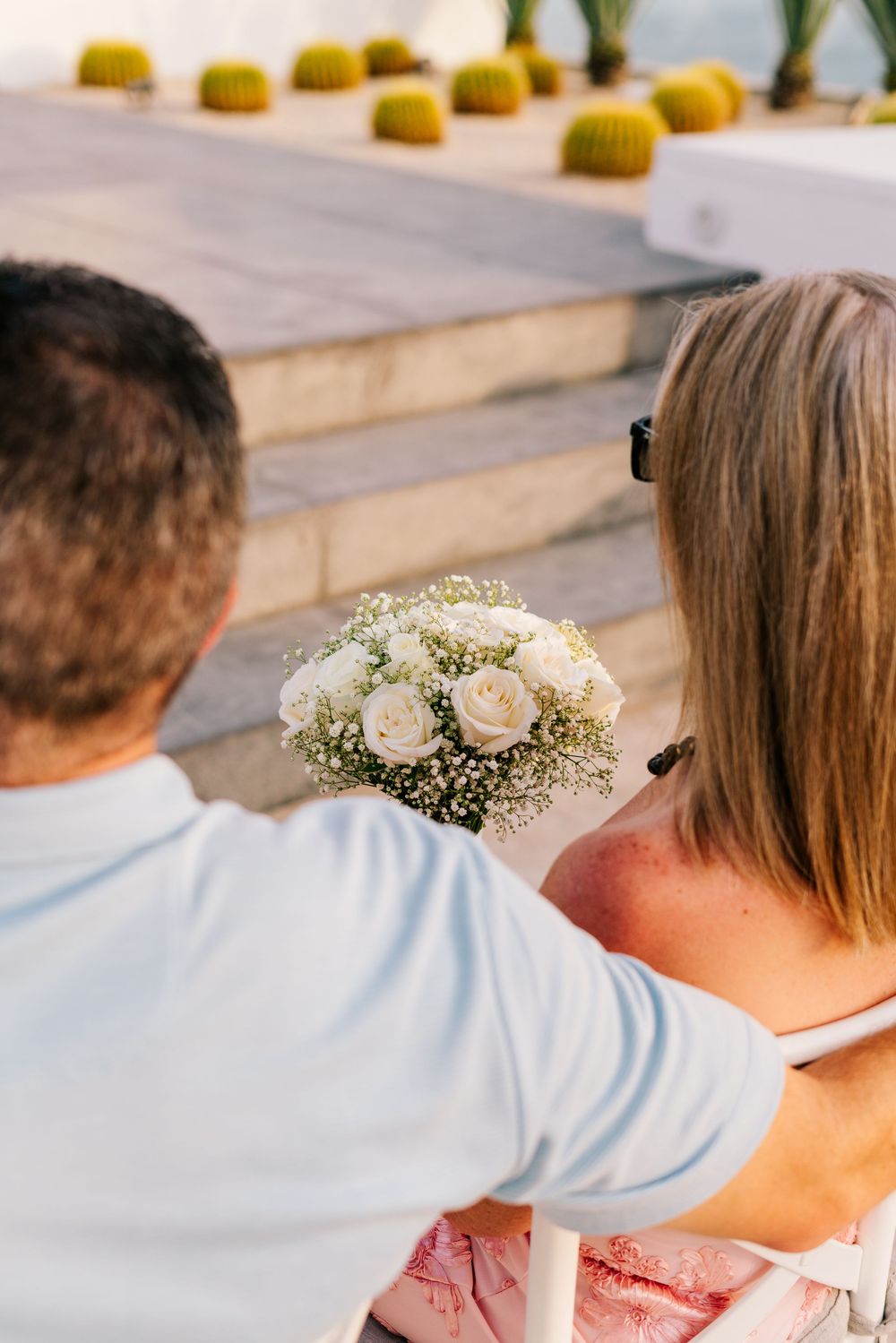 Couple viewed from behind, man presenting white rose bouquet with baby's breath to woman on sunny outdoor steps.