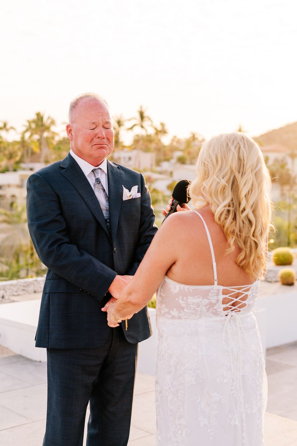 Emotional groom in navy suit holds hands with blonde bride in lace wedding dress during outdoor ceremony.