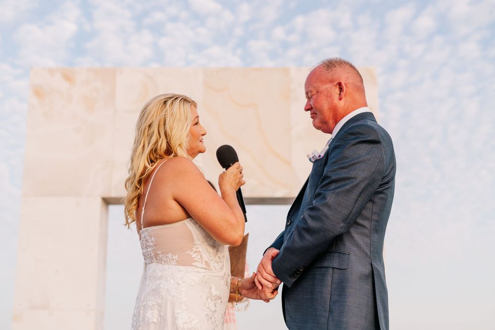 Bride reading vows to groom at outdoor wedding ceremony with large stone cross backdrop at sunset.