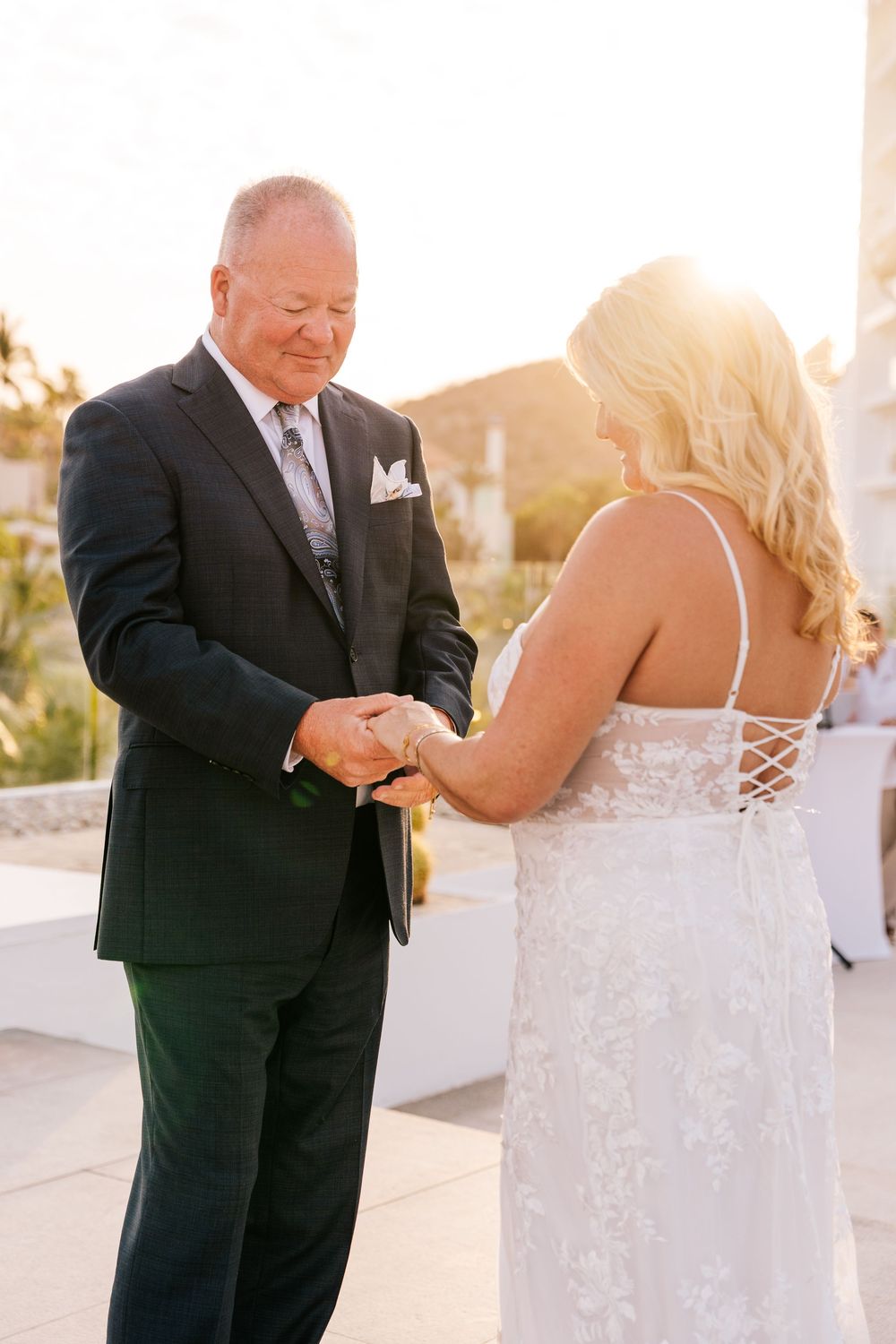 A groom in a black suit places a wedding ring on his bride in a lace dress during a golden hour outdoor ceremony.