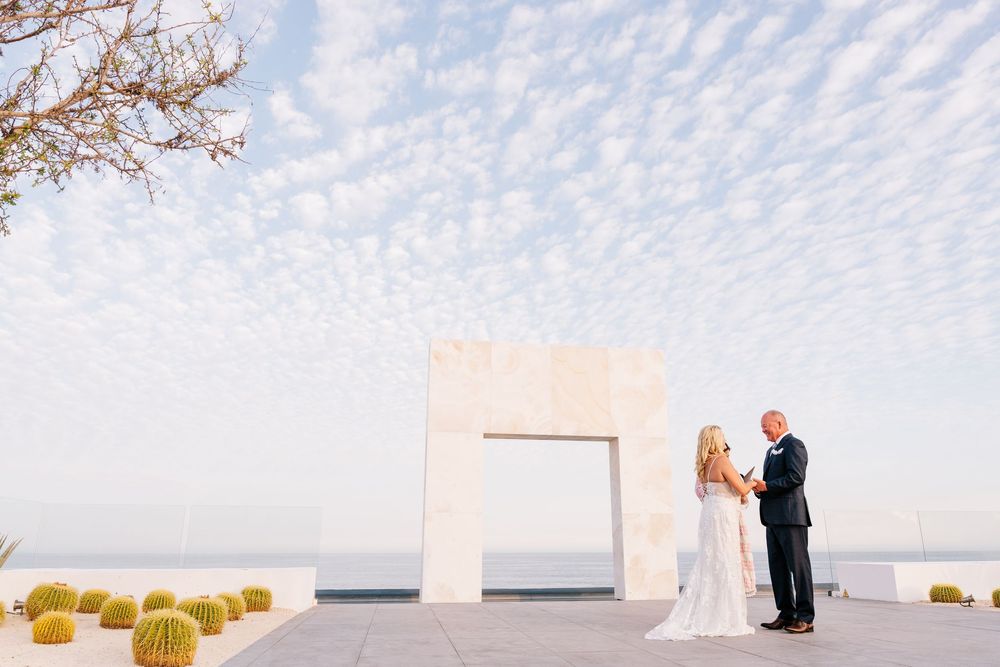 Couple exchanges vows under a white arch at an outdoor wedding ceremony with cacti and ocean views.