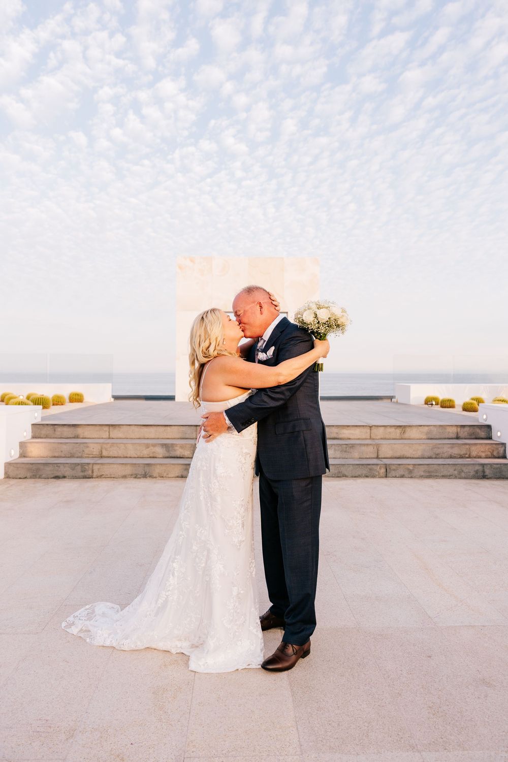 Bride and groom share first kiss at outdoor wedding ceremony with ocean view and cloudy sky backdrop.