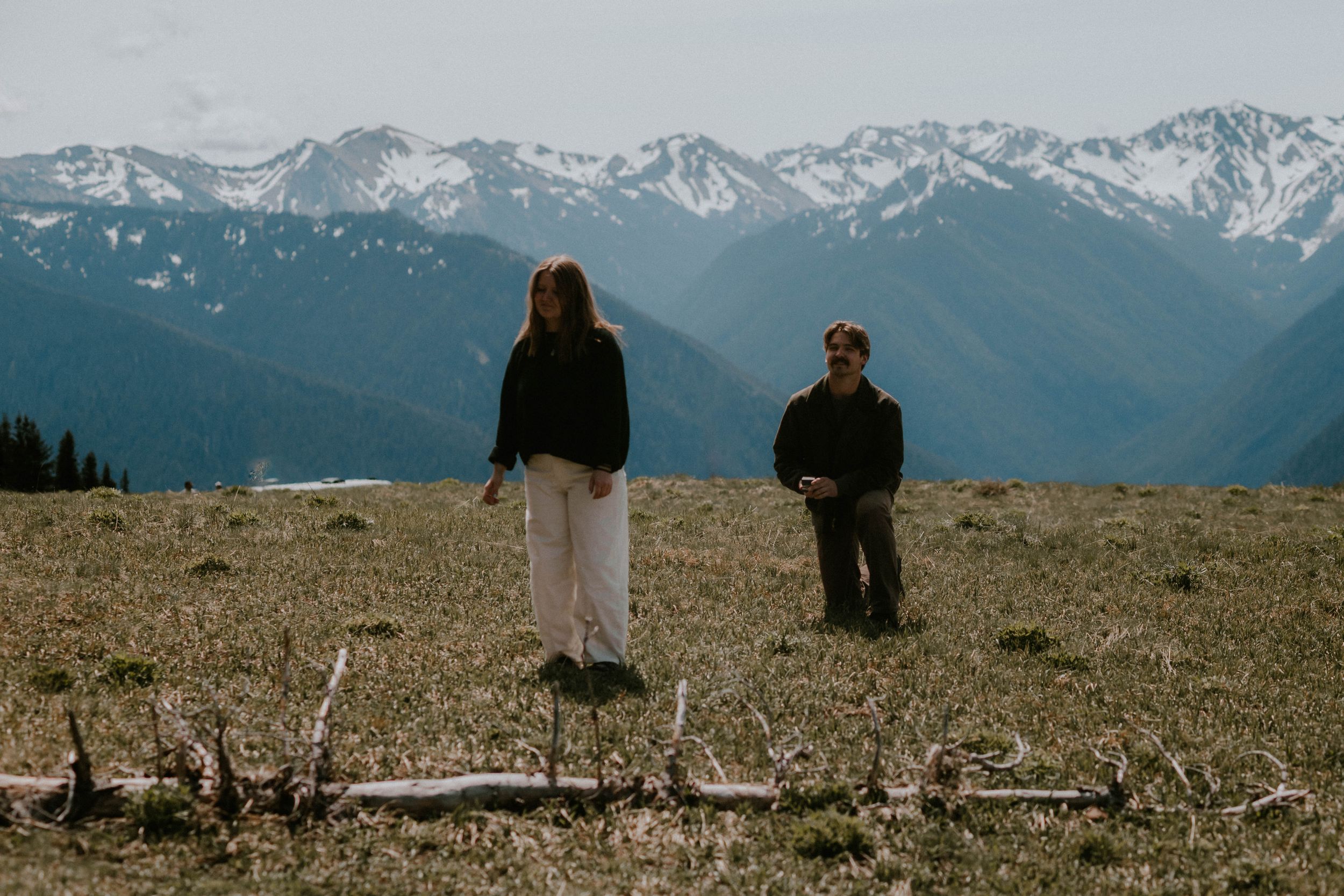 Two people stand in a mountain meadow with snow-capped peaks in the background on a clear day.