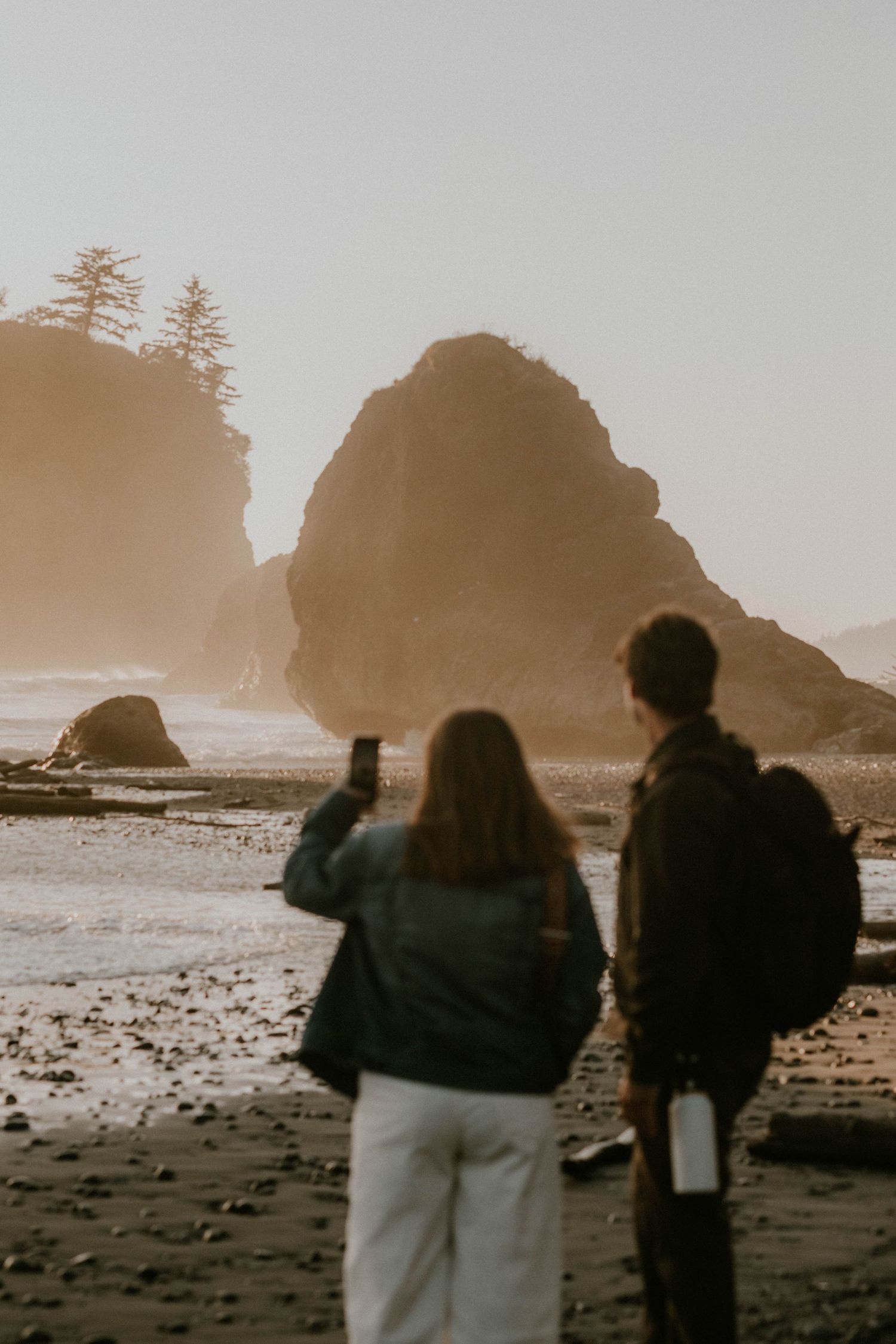 Two hikers stand on a misty beach photographing dramatic sea stacks at golden hour on the Pacific Northwest coast.