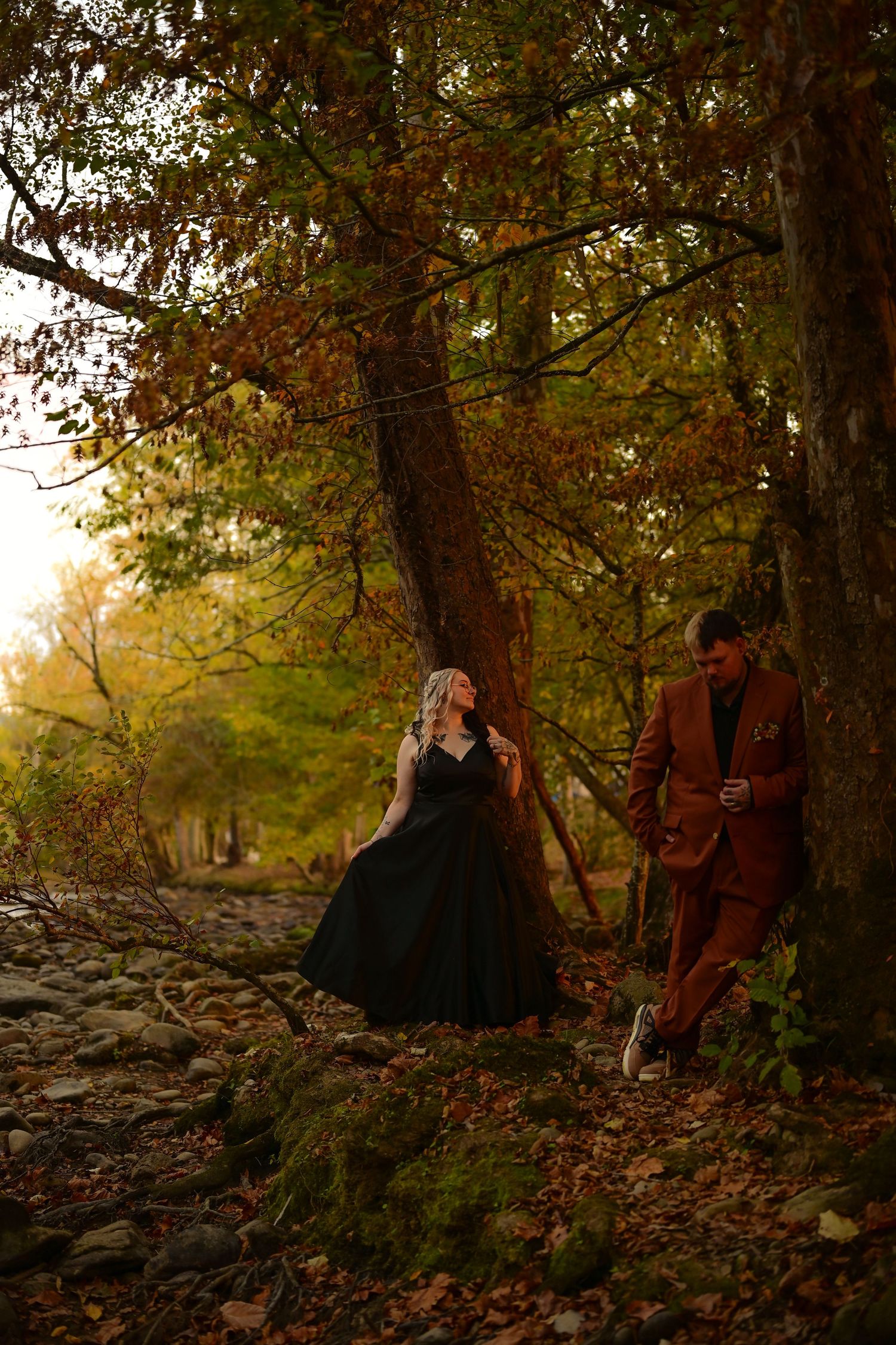 Couple in autumn forest, woman in black gown leaning on tree, man in rust suit, fall foliage backdrop.