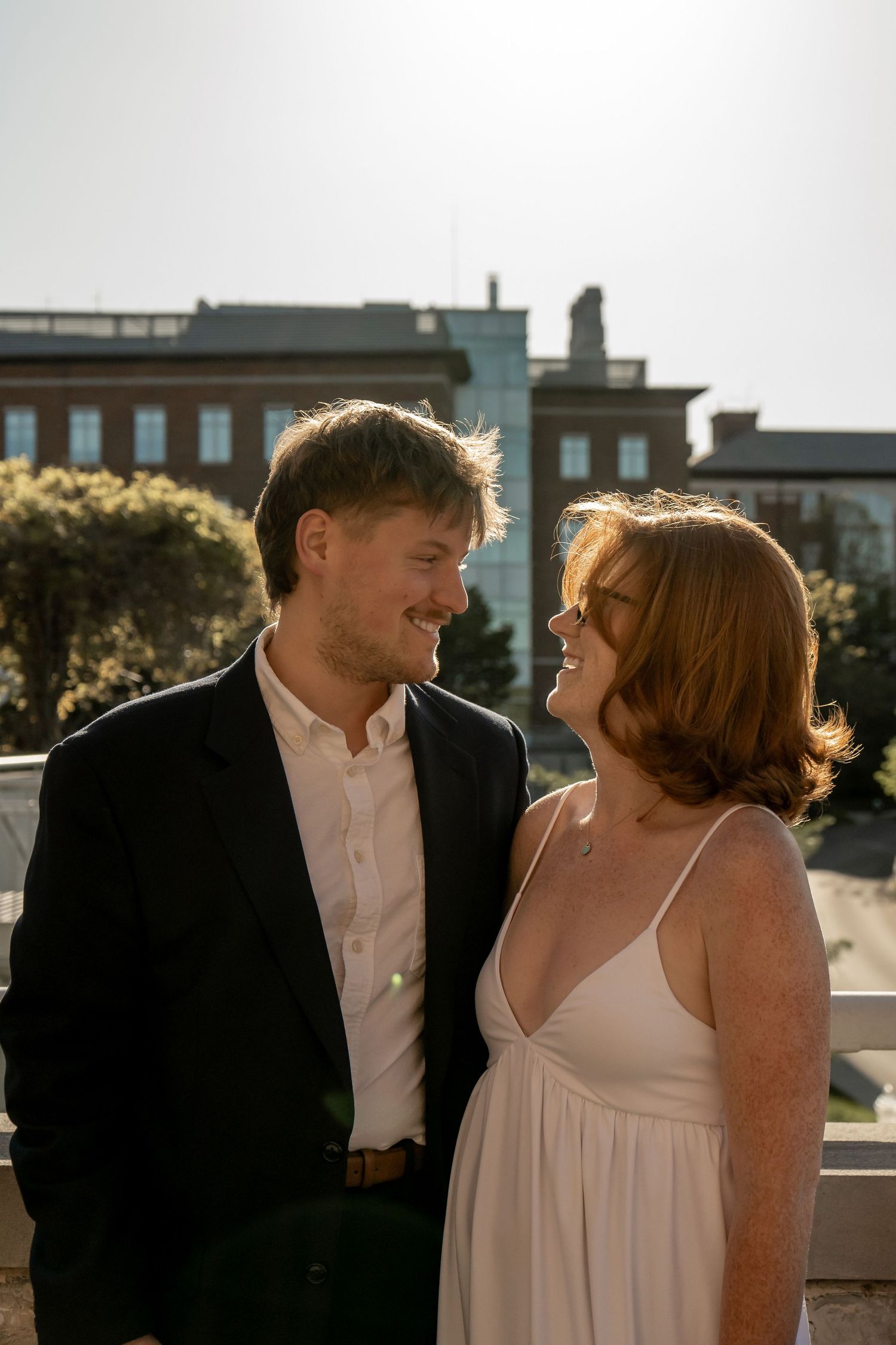 Young couple smiling at each other outdoors in sunlight near historic brick building.