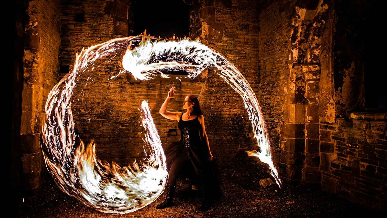 Fire performer creates glowing circular light trails in dark stone ruins at night.